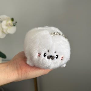 May include: A white plush seal toy with a silver chain. The seal has black eyes, a black nose, and pink blush marks on its cheeks. The toy is round and fluffy.