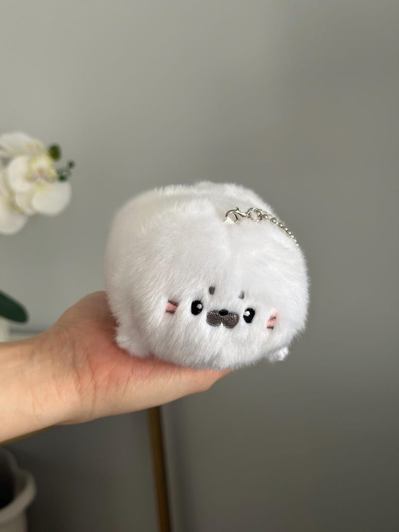 May include: A white plush seal toy with a silver chain. The seal has black eyes, a black nose, and pink blush marks on its cheeks. The toy is round and fluffy.