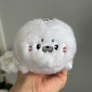 May include: A white plush seal keyring with embroidered features. The seal has black eyes, a grey nose, and pink blush marks. It is round and fluffy, with a silver keyring attachment.