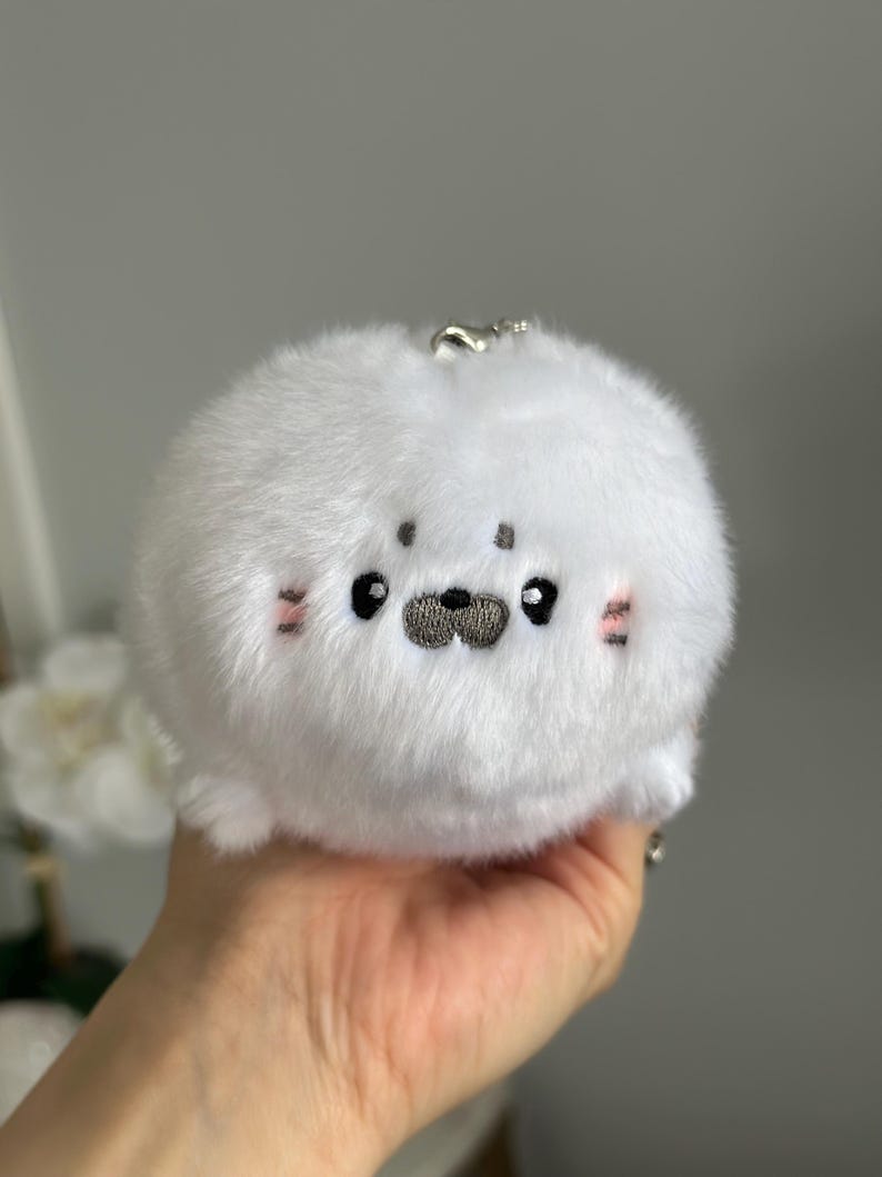May include: A white plush seal keyring with embroidered features. The seal has black eyes, a grey nose, and pink blush marks. It is round and fluffy, with a silver keyring attachment.