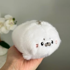 May include: A white plush seal keyring with a silver clasp. The seal has black eyes, a grey nose, and pink blush marks. The plush toy is soft and fluffy.