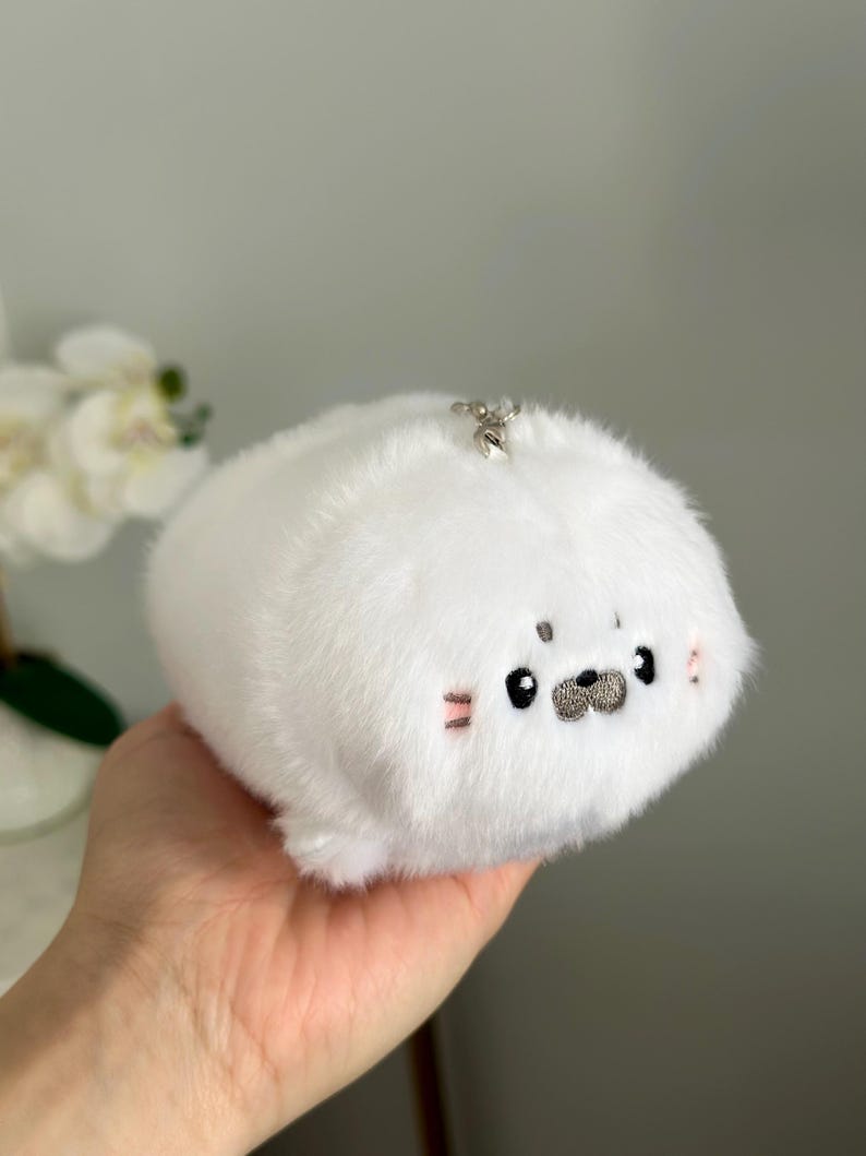 May include: A white plush seal keyring with a silver clasp. The seal has black eyes, a grey nose, and pink blush marks. The plush toy is soft and fluffy.