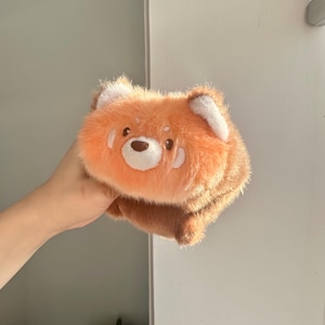 May include: A plush, orange red panda stuffed animal with white accents on the ears and a white muzzle. The stuffed animal has a brown nose and small, embroidered eyes. The plush toy is held up in front of a white door.