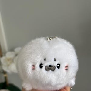 May include: A soft, white plush seal keyring. The seal features embroidered black eyes, a grey nose, and pink blush marks on its cheeks. A silver metal clasp is attached.