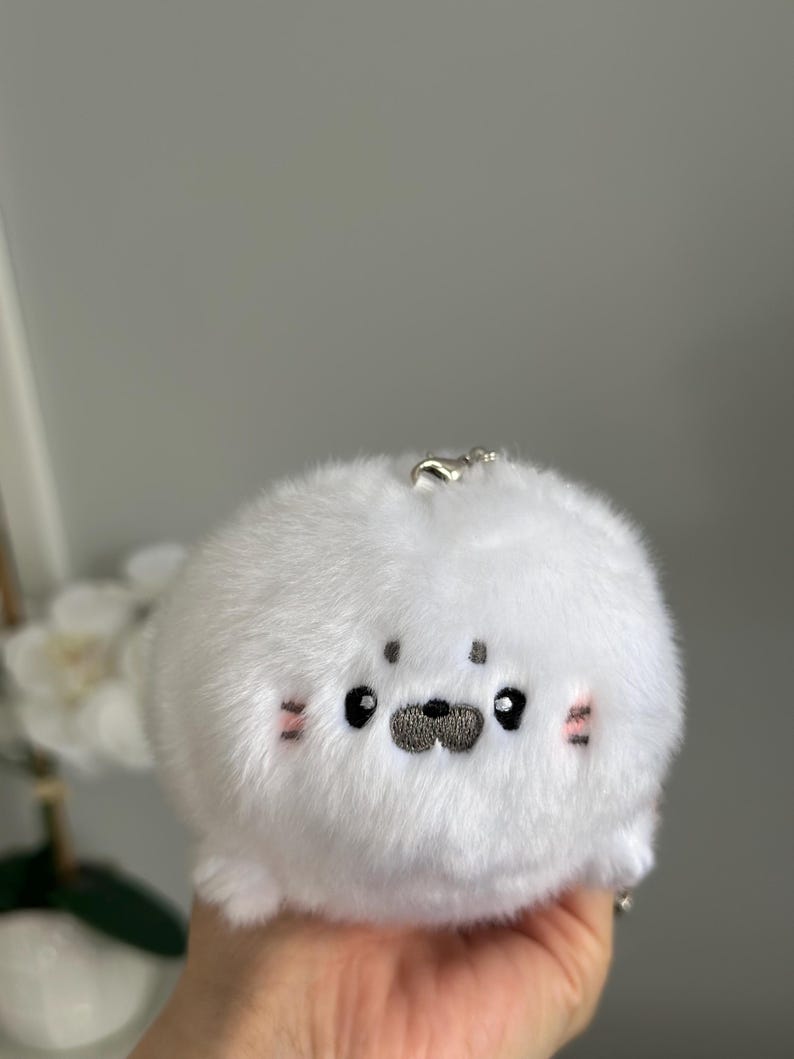 May include: A soft, white plush seal keyring. The seal features embroidered black eyes, a grey nose, and pink blush marks on its cheeks. A silver metal clasp is attached.