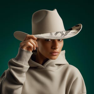 May include: A light gray cowboy hat with a decorative brim featuring a black and white horse scene. The hat is held up by a person wearing a light gray hoodie. The background is a dark teal color.