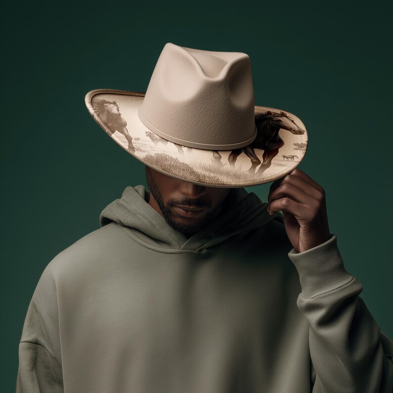 May include: A beige cowboy hat with a detailed illustration of running horses on the brim. The hat is worn over a sage green hoodie. The background is a solid, dark teal color.