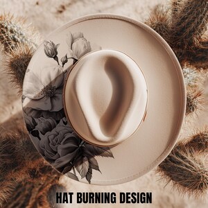 Op de afbeelding: Een crèmekleurige fedora-stijl hoed met een bloemenontwerp in zwart en wit. De hoed heeft een brede rand en een afgeronde kroon. De tekst "HAT BURNING DESIGN" staat onderaan de afbeelding. De hoed is omgeven door cactussen.