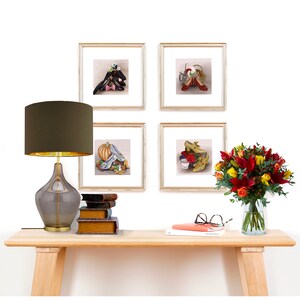 May include: Four framed illustrations of shoes from classic fairy tales, including Cinderella's glass slipper, Dorothy's ruby slippers, and the red shoes from the story of the red shoes. The illustrations are displayed on a white wall above a wooden console table with a lamp, books, and a vase of flowers.