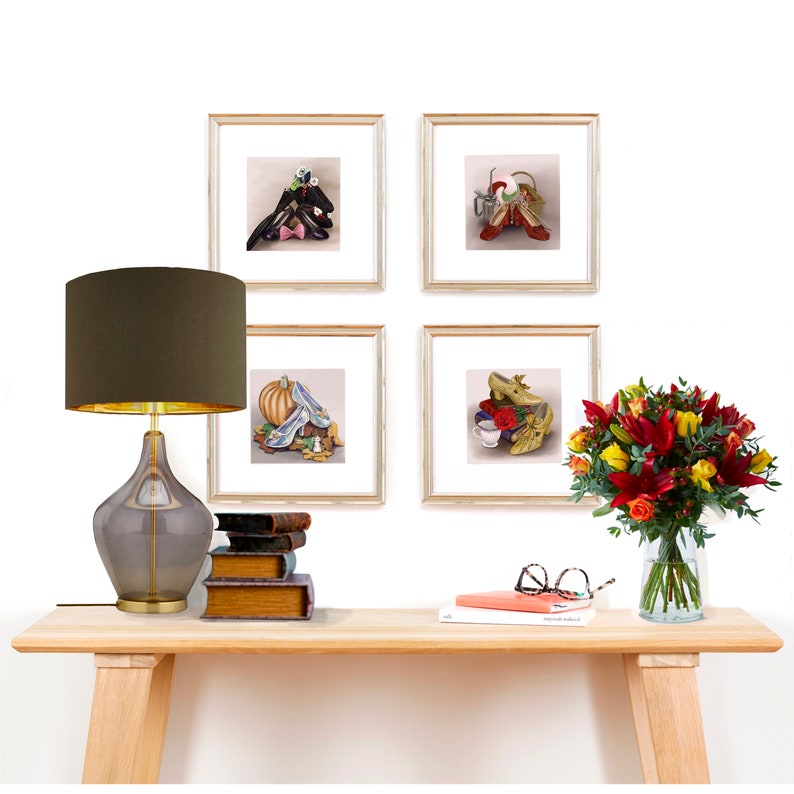 May include: Four framed illustrations of shoes from classic fairy tales, including Cinderella's glass slipper, Dorothy's ruby slippers, and the red shoes from the story of the red shoes. The illustrations are displayed on a white wall above a wooden console table with a lamp, books, and a vase of flowers.