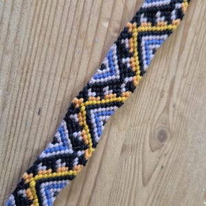 Può includere: Un braccialetto di amicizia intrecciato con un motivo geometrico in nero, blu, giallo e arancione.