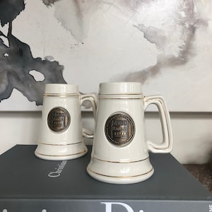 May include: Two off-white ceramic beer steins with gold trim and handles. Each stein features a circular emblem with the text "Hamm's 100 Years". The steins are displayed on a dark gray book.