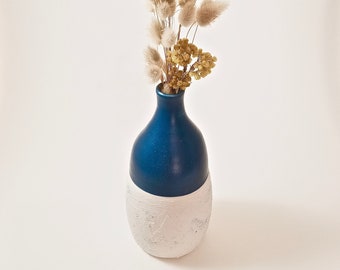May include: A two-toned vase with a smooth, deep blue upper section and a textured, matte white lower base. The vase contains a dried floral arrangement featuring fluffy beige bunny tail grass and small clusters of yellow dried flowers.