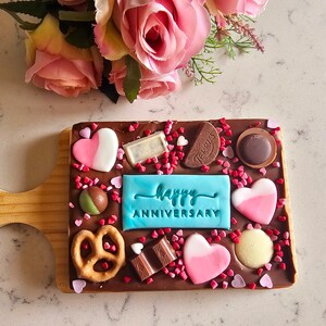 Peut inclure: Une barre de chocolat avec un message glac&eacute; bleu qui dit "Joyeux Anniversaire" sur une planche en bois. La barre de chocolat est d&eacute;cor&eacute;e de c&oelig;urs roses et blancs, de bretzels et de p&eacute;pites.