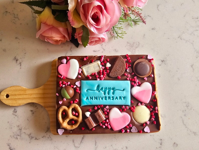 Peut inclure: Une barre de chocolat avec un message glac&eacute; bleu qui dit "Joyeux Anniversaire" sur une planche en bois. La barre de chocolat est d&eacute;cor&eacute;e de c&oelig;urs roses et blancs, de bretzels et de p&eacute;pites.