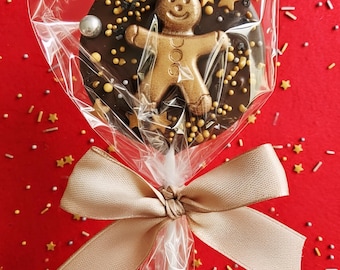 Puede incluir: Una piruleta de chocolate con un hombre de jengibre dorado, estrellas doradas y chispas. La piruleta está envuelta en celofán transparente y atada con una cinta dorada. El fondo es rojo con chispas doradas en forma de estrella.