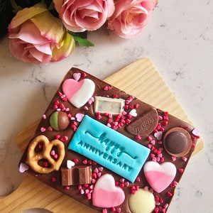 Peut inclure: Une barre de chocolat avec un message "Joyeux Anniversaire" bleu et blanc, entour&eacute;e de c&oelig;urs roses et blancs, de bretzels et de bonbons au chocolat.