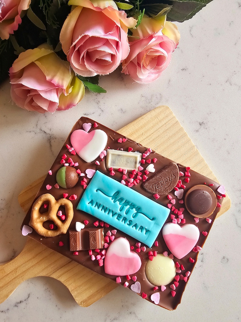 Peut inclure: Une barre de chocolat avec un message "Joyeux Anniversaire" bleu et blanc, entour&eacute;e de c&oelig;urs roses et blancs, de bretzels et de bonbons au chocolat.