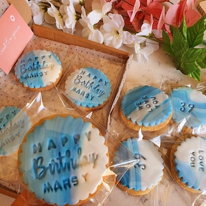 May include: Round sugar biscuits with scalloped edges, featuring a blue and white marbled icing finish. The biscuits are individually wrapped in clear plastic and embossed with the text Happy Birthday Marsy, 39, or We Love You in a dark blue font.