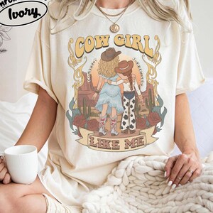 May include: An ivory short-sleeve t-shirt featuring a graphic print of two figures in cowboy hats standing in a desert landscape with cacti and roses. The text reads COW GIRL LIKE ME. A Comfort Colors Ivory label is visible in the upper left corner.