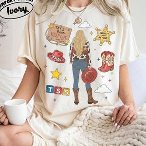 May include: An ivory short-sleeve t-shirt featuring a graphic print of a person with long blonde hair, wearing a cow-print jacket, blue jeans, and brown boots. The design includes a wooden sign reading Tay's Room Keep Out, a sheriff star, red cowboy hats, and blocks labelled TS5.