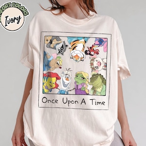 May include: Ivory-colored t-shirt featuring a framed graphic of cartoon characters, including a raccoon, tiger, and frog. The graphic is labeled "Once Upon A Time". The shirt has short sleeves and a crew neck.