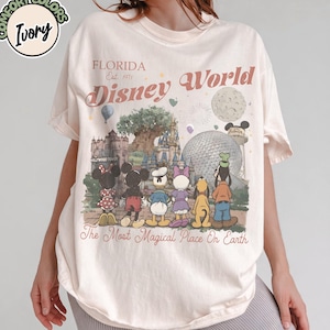 May include: Ivory-colored t-shirt with a Disney World graphic. The design features Disney characters in front of park landmarks, with text that reads "Florida Est. 1971 Disney World" and "The Most Magical Place On Earth."