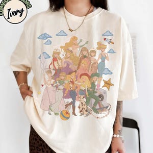May include: An ivory t-shirt featuring a graphic print of multiple animated characters in various outfits, including a space suit, cowgirl attire, and dresses. The design includes blue clouds, a small rocket, a star, and a ball. Text reads COMFORT COLORS IVORY.