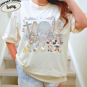 May include: Ivory-colored t-shirt with a colorful graphic featuring cartoon characters and landmarks. The graphic includes Mickey Mouse, Donald Duck, and other characters, along with the Eiffel Tower and other world landmarks. The text "COMFORT COLORS" and "Ivory" are printed on the shirt.