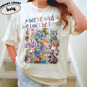 May include: Ivory-colored t-shirt with a colorful graphic featuring characters from Zootopia and the text "We're Wild We Can't Be Tamed." The shirt is a Comfort Colors brand.