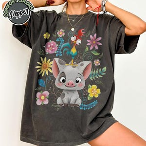 May include: Dark gray t-shirt featuring a cartoon pig with a rooster on its head, surrounded by colorful flowers and foliage. The shirt has the text "Comfort Colors Pepper" in a circle on the upper left.