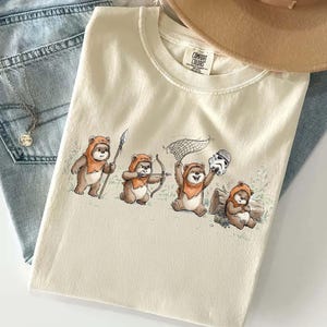 Puede incluir: Camiseta Comfort Colors color crema con una ilustración de cuatro Ewoks. Uno sostiene una lanza, otro un arco y flechas, un tercero una red y el último un casco de Stormtrooper. La camiseta está doblada sobre unos vaqueros.