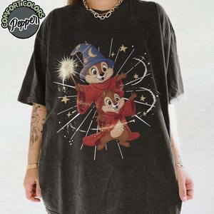 May include: Dark gray t-shirt featuring a graphic of two cartoon chipmunks dressed as wizards, one holding a wand. The chipmunks wear red robes and wizard hats, with a background of stars and lines. The shirt has the text "Comfort Colors Pepper" in a circular design.