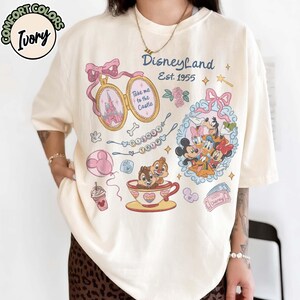Peut inclure: T-shirt ivoire avec des graphismes de Disneyland. Le design comprend un m&eacute;daillon avec un ch&acirc;teau, Mickey Mouse et ses amis, Tic et Tac dans une tasse &agrave; th&eacute;, et le texte "Disneyland Est. 1955".