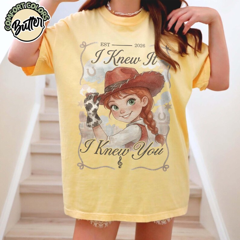 Peut inclure: Un t-shirt jaune p&acirc;le avec une illustration de cow-girl aux cheveux roux tress&eacute;s et un chapeau de cowboy marron. Le texte indique EST 2026, I Knew It, et I Knew You, avec une note de musique et une bordure en lasso. Un logo Comfort Colors Butter est pr&eacute;sent.