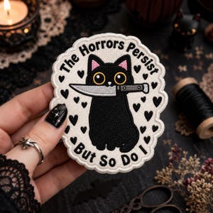 May include: A circular embroidered patch with a scalloped edge featuring a black cat holding a silver knife in its mouth. The background is off-white with small black hearts. The text reads, The Horrors Persist But So Do I in black letters.