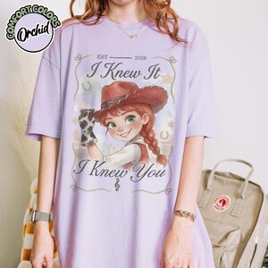 Peut inclure: Un t-shirt &agrave; manches courtes violet clair avec une illustration de personnage portant un chapeau de cow-boy marron et un gant &agrave; motif vache. Le texte indique "EST 2026 I Knew It I Knew You" avec une note de musique, des &eacute;toiles et des fers &agrave; cheval.