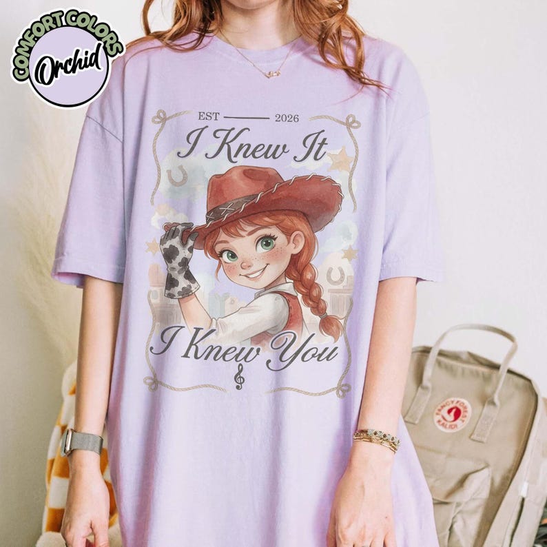 Peut inclure: Un t-shirt &agrave; manches courtes violet clair avec une illustration de personnage portant un chapeau de cow-boy marron et un gant &agrave; motif vache. Le texte indique "EST 2026 I Knew It I Knew You" avec une note de musique, des &eacute;toiles et des fers &agrave; cheval.