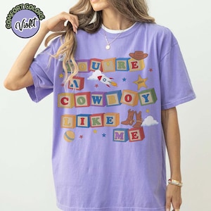 May include: A violet short-sleeve t-shirt featuring a graphic of colorful alphabet blocks spelling out the text "YOU'RE A COWBOY LIKE ME". The design includes illustrations of a rocket ship, a sheriff badge, a cowboy hat, and brown boots.