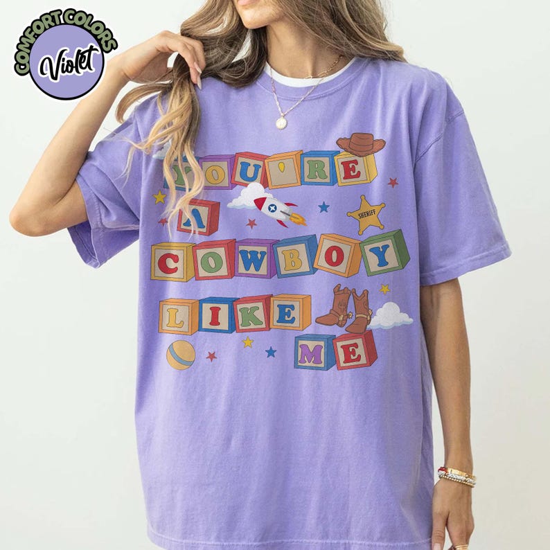 May include: A violet short-sleeve t-shirt featuring a graphic of colorful alphabet blocks spelling out the text "YOU'RE A COWBOY LIKE ME". The design includes illustrations of a rocket ship, a sheriff badge, a cowboy hat, and brown boots.
