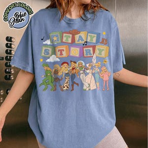 May include: A blue short-sleeve t-shirt featuring a graphic print of colourful alphabet blocks spelling TAY STORY above a line of illustrated characters dressed in various outfits, including a green dinosaur suit and western-style clothing.
