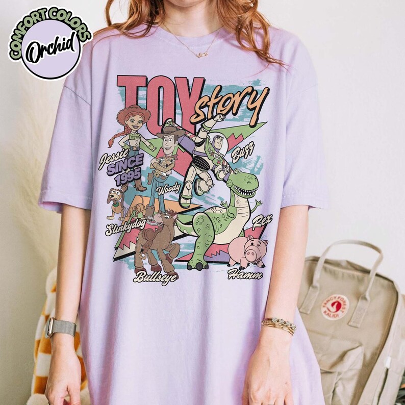 May include: A light purple t-shirt featuring the words "TOY STORY" in a retro font, with images of characters like Woody, Buzz Lightyear, and Rex. The shirt also includes the text "Jessie Since 1995".
