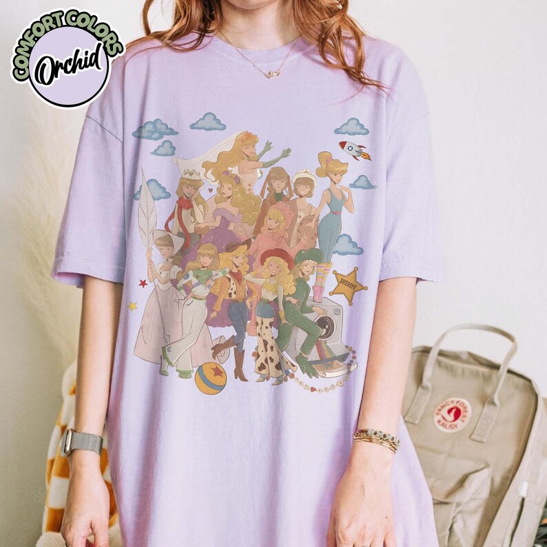 May include: A light purple short-sleeve t-shirt featuring a graphic print of multiple animated characters in various outfits, including a cowboy hat, space suit, and dresses, alongside clouds, a rocket, a ball, and a sheriff star badge.