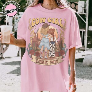 May include: A light pink t-shirt featuring a graphic illustration of two figures in cowboy hats standing in a desert landscape with cacti and roses. The text reads COW GIRL and LIKE ME, with the phrase I KNOW I KNOW YOU printed in a circular arc.