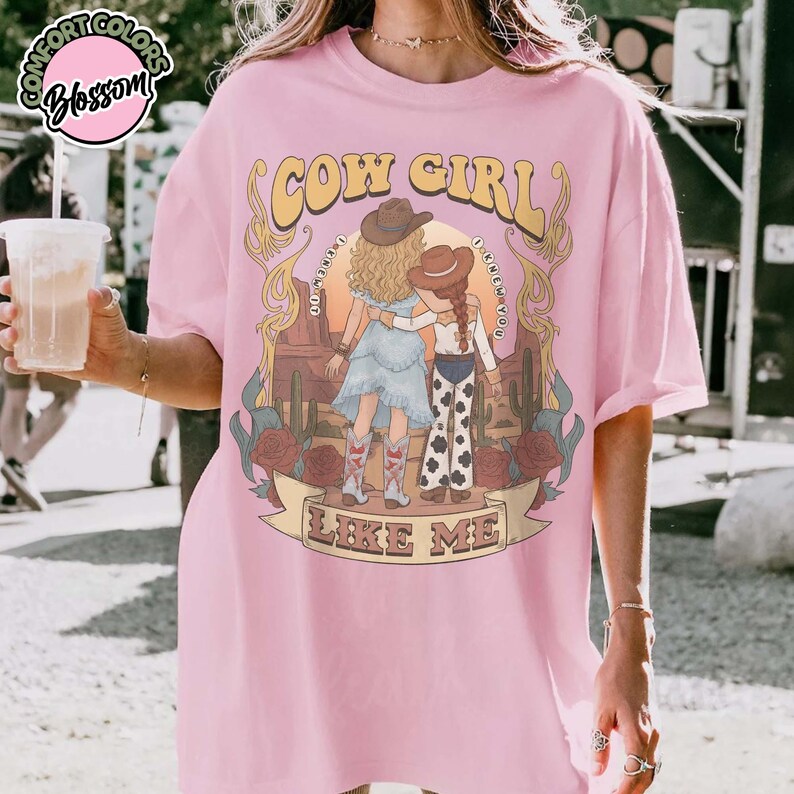 May include: A light pink t-shirt featuring a graphic illustration of two figures in cowboy hats standing in a desert landscape with cacti and roses. The text reads COW GIRL and LIKE ME, with the phrase I KNOW I KNOW YOU printed in a circular arc.