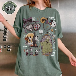 May include: Sage green t-shirt featuring a graphic design with characters and elements from "The Nightmare Before Christmas." The design includes Jack Skellington, Sally, Oogie Boogie, and other spooky elements. The shirt has a relaxed fit.