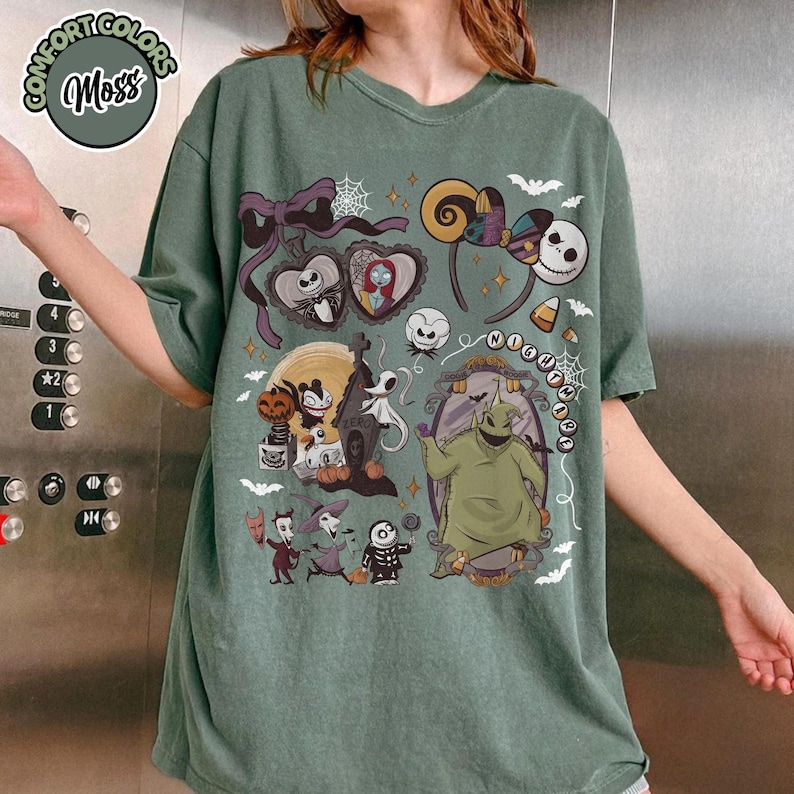 May include: Sage green t-shirt featuring a graphic design with characters and elements from "The Nightmare Before Christmas." The design includes Jack Skellington, Sally, Oogie Boogie, and other spooky elements. The shirt has a relaxed fit.
