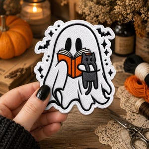 Puede incluir: Un parche bordado que muestra un fantasma blanco con ojos negros sosteniendo un libro naranja abierto y un pequeño gato negro. El diseño incluye contornos negros, pequeños murciélagos negros y formas de estrellas alrededor del fantasma sobre tela blanca.