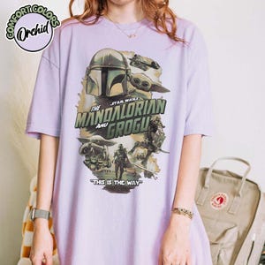 May include: A light purple t-shirt featuring a graphic print of The Mandalorian and Grogu from Star Wars. The design includes the text "The Mandalorian and Grogu" and "This is the Way" with illustrations of the characters and a vehicle.