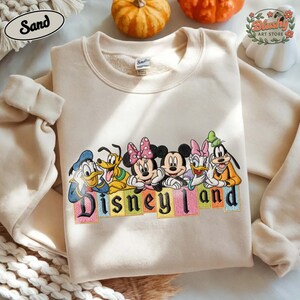 May include: A sand-colored crewneck sweatshirt featuring an embroidered graphic of Disney characters including Donald Duck, Pluto, Minnie Mouse, Mickey Mouse, Daisy Duck, and Goofy. Below the characters, the word Disneyland is stitched in black letters.
